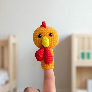 May include: A handmade, crocheted finger puppet shaped like a rooster. The puppet is primarily orange with a red comb and wattle, and a yellow beak. The puppet is on a finger.