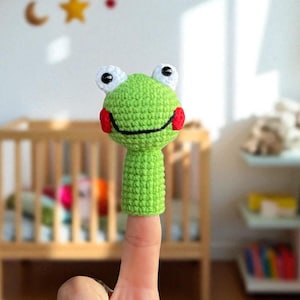 Puede incluir: Una marioneta de dedo de rana verde hecha a mano con ojos blancos y mejillas rojas. La marioneta está sujeta por un dedo. En el fondo hay una cuna de madera y un móvil con una luna y estrellas.