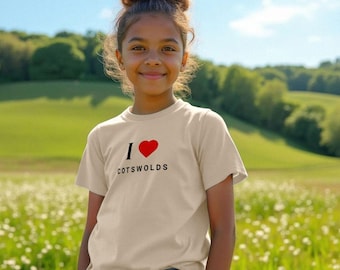 Puede incluir: Una persona joven con una camiseta beige con el texto "I ❤️ COTSWOLDS" en negro y rojo. La persona sonríe y está de pie en un campo de flores con una colina verde y cielo azul al fondo.