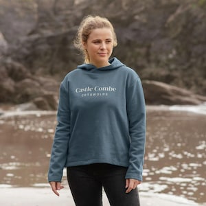 May include: A teal hoodie with the text "Castle Combe Cotswolds" in white. The hoodie has a hood and long sleeves. The person is wearing black trousers. The hoodie is made of a soft material.