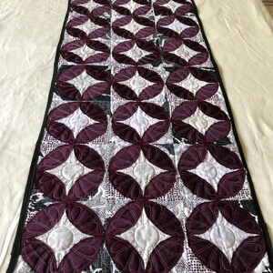 May include: A purple and white quilted table runner with a geometric pattern. The runner has a black border.