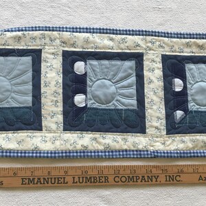 May include: A quilted table runner featuring three square designs with a sunburst pattern in shades of blue. The runner has a cream background with blue floral accents and a blue gingham border. A wooden ruler is at the bottom.