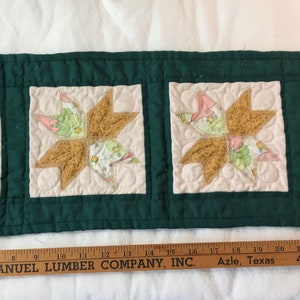 May include: A green quilted table runner with four beige squares featuring a brown and white star design. The runner is 28 inches long and has a wooden ruler with the text "EMANUEL LUMBER COMPANY, INC. Azle, Texas 444-2566" printed on it.