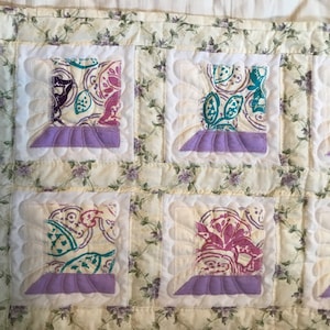 May include: A quilt with a floral pattern and a patchwork design. The quilt is made up of squares with different patterns and colors, including purple, pink, and green. The quilt is edged with a white border.