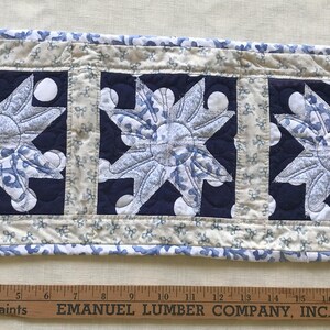 May include: A quilted table runner with three navy blue squares, each featuring a light blue floral design. The runner has a cream-colored border with blue floral accents. A wooden ruler is at the bottom.