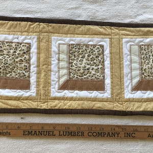 May include: A rectangular, handmade quilted table runner. It features three square panels with a leopard print design, framed by white and brown accents. The border is gold with a dark brown trim.