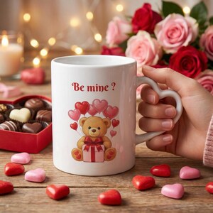 Può includere: Tazza in ceramica bianca con manico a forma di cuore, con un orsacchiotto che tiene un regalo e palloncini a forma di cuore. La tazza ha il testo "Be mine ?" ed è circondata da cioccolatini e rose.