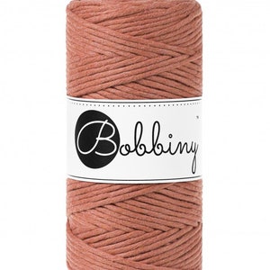 K&ouml;nnte beinhalten: Eine Spule aus staubrosa Makramee-Kordel. Die Kordel ist eng gewickelt. Ein wei&szlig;es Etikett mit dem Wort "Bobbiny" in einem schwarzen Kreis ist um die Mitte der Spule gewickelt. Geeignet f&uuml;r verschiedene Bastelprojekte.
