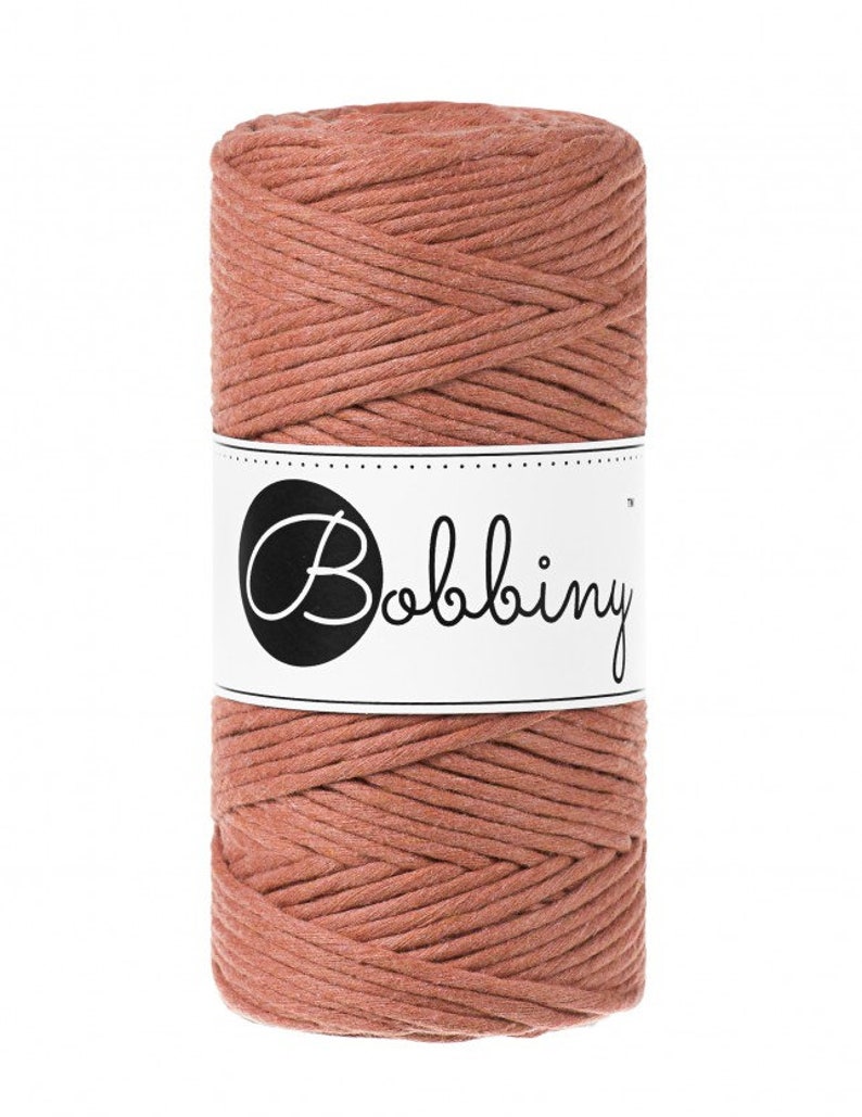 K&ouml;nnte beinhalten: Eine Spule aus staubrosa Makramee-Kordel. Die Kordel ist eng gewickelt. Ein wei&szlig;es Etikett mit dem Wort "Bobbiny" in einem schwarzen Kreis ist um die Mitte der Spule gewickelt. Geeignet f&uuml;r verschiedene Bastelprojekte.