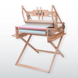 May include: A wooden weaving loom with a colorful woven textile in pink, blue, and white. The loom is on a folding stand, and the frame is made of light-colored wood. The loom is designed for creating woven fabrics and textiles.