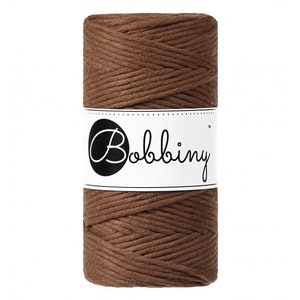 K&ouml;nnte beinhalten: Eine Spule braunes Makramee-Garn mit einem wei&szlig;en Etikett mit dem Wort "Bobbiny" in schwarzer, stilisierter Schrift. Das Garn ist eng gewickelt und zeigt eine strukturierte Oberfl&auml;che. Ideal f&uuml;r Bastelprojekte.