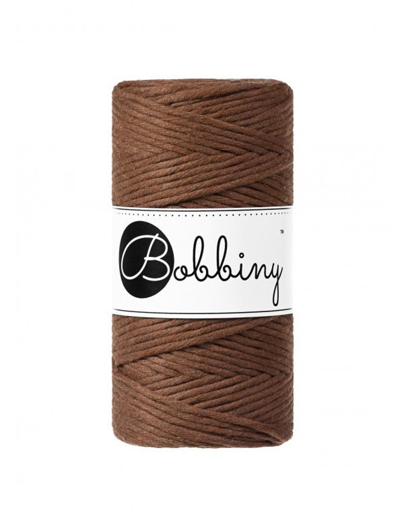 K&ouml;nnte beinhalten: Eine Spule braunes Makramee-Garn mit einem wei&szlig;en Etikett mit dem Wort "Bobbiny" in schwarzer, stilisierter Schrift. Das Garn ist eng gewickelt und zeigt eine strukturierte Oberfl&auml;che. Ideal f&uuml;r Bastelprojekte.