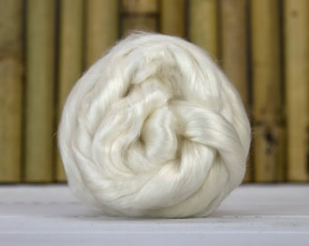 May include: A tightly wound ball of off-white fiber, likely wool or silk, is the focal point. The fibers have a soft, lustrous appearance, suggesting a high-quality material. The background features vertical bamboo stalks.