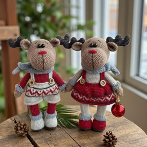 May include: Two handmade crochet reindeer dolls. The reindeer have brown bodies, black antlers, and wear red and white outfits with blue scarves. One doll holds a red ornament. The dolls are on a wooden surface.