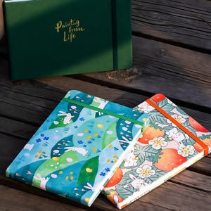 May include: A collection of notebooks with colorful covers. One notebook is dark green with the text "Painting from Life" in gold. Two notebooks have illustrated covers with bunnies and strawberries. The notebooks are on a wooden surface.