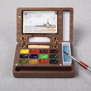 以下が含まれることがあります： 濃い木製の水彩絵の具ボックス。様々な色の絵の具が入った12個の長方形の仕切り、小さな長方形のスポンジ、白いミキシングトレイ、灯台の水彩画が収められています。木製の柄の絵筆が付属しています。
