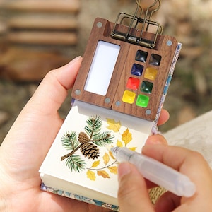 Op de afbeelding: Een houten aquarelpalet en notitieblokset. Het palet heeft een rechthoekige witte ruimte en een selectie van kleurrijke verven. Het notitieblok bevat een botanische illustratie van dennennaalden, een dennenappel en herfstbladeren. Een heldere waterkwast wordt boven het notitieblok gehouden.