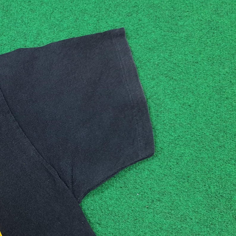 May include: A black short-sleeved t-shirt featuring a visible hemmed sleeve edge and a smooth fabric texture. The garment is displayed flat, showcasing the dark solid color of the textile and the clean stitching along the sleeve seam and shoulder area.