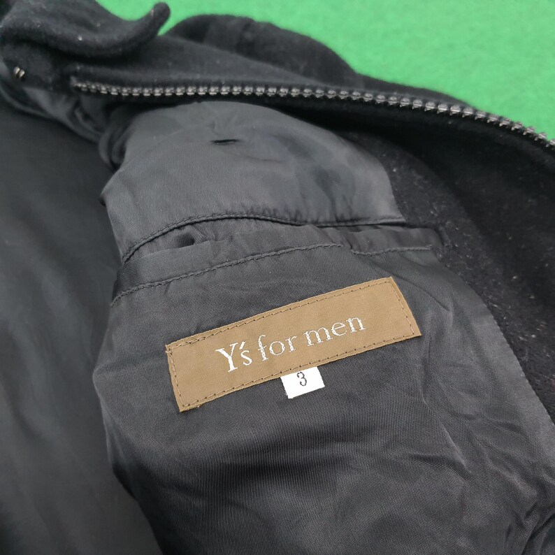 K&ouml;nnte beinhalten: Nahaufnahme des Innenraums einer schwarzen Jacke. Das Futter ist dunkel mit einer Tasche und einem Etikett mit der Aufschrift "Y's for men". Ein Gr&ouml;&szlig;enetikett mit der Nummer 3 ist ebenfalls sichtbar. Die Jacke ist aus strukturiertem Stoff.