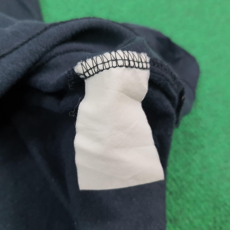 May include: A dark navy blue garment featuring a blank white rectangular fabric care label attached to the interior neckline with black stitching. The fabric appears to have a soft, brushed texture, and the label is empty with no visible text.