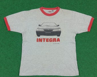 May include: A grey t-shirt with a red collar and sleeve trim. The front displays a black car graphic and the red word "INTEGRA" underneath. The shirt is set against a green backdrop.