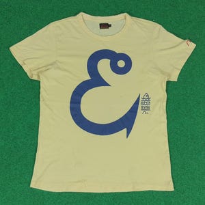 Puede incluir: Una camiseta de manga corta de color amarillo pálido con un gran gráfico de ampersand azul oscuro en la parte delantera. A la derecha del ampersand hay un pequeño logotipo con un icono de pez, texto en japonés y la palabra EVISU. Un pequeño logotipo rojo está en la manga izquierda.