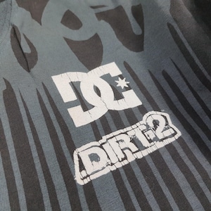 May include: A dark gray t-shirt featuring a black flame-like pattern and a white graphic print. The print displays the DC logo above the text DIRT 2, with visible cracking in the white ink. The fabric has a ribbed texture and a crew neckline.