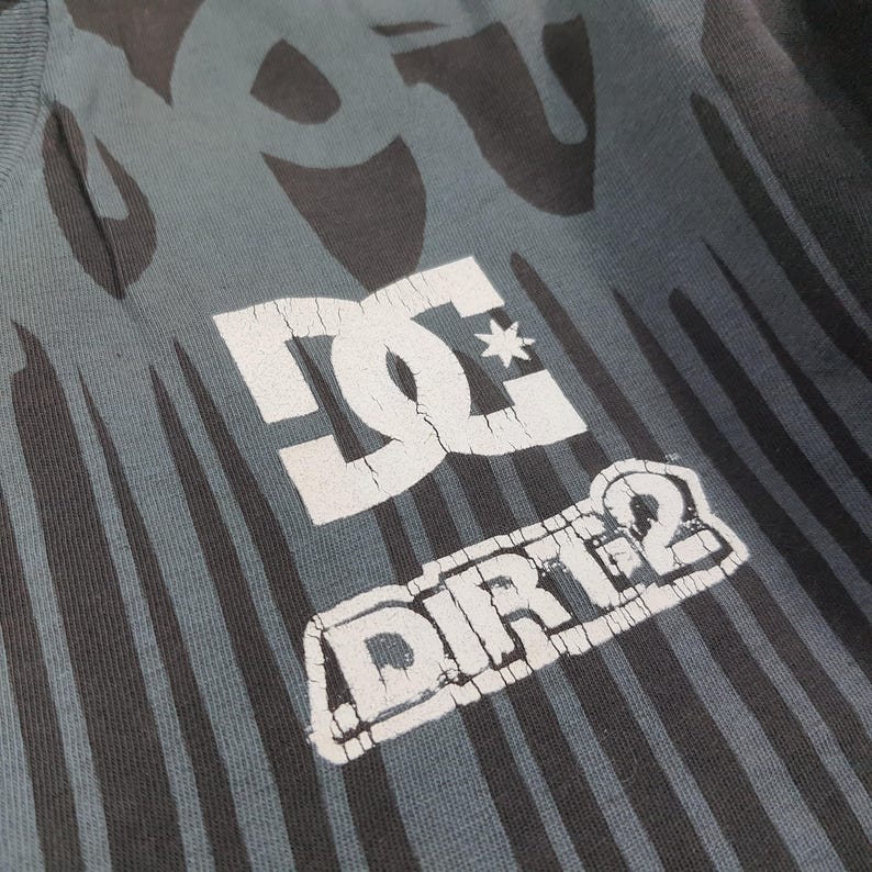 May include: A dark gray t-shirt featuring a black flame-like pattern and a white graphic print. The print displays the DC logo above the text DIRT 2, with visible cracking in the white ink. The fabric has a ribbed texture and a crew neckline.