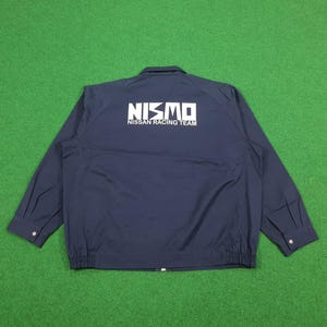Puede incluir: Una chaqueta azul marino de manga larga con un estampado gráfico blanco en la espalda que dice NISMO NISSAN RACING TEAM. La chaqueta tiene un cuello clásico, una cremallera frontal completa y puños abotonados con un dobladillo elástico.