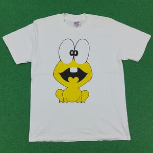 Op de afbeelding: Een wit t-shirt met korte mouwen en een grote, gecentreerde afbeelding van een gele cartoonkikker met extra grote witte ogen, een zwarte mond en één witte tand. Het shirt heeft een ronde hals en een Hanes-label in de kraag.