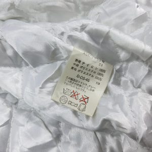 May include: Close-up of a white quilted fabric with a tag. The tag has Japanese text and reads "ASK-2653W 23". The tag also includes fabric composition details: 100% nylon, 100% polyester, and 100% polyester. Additional text on the tag includes "D-OS-46 118".