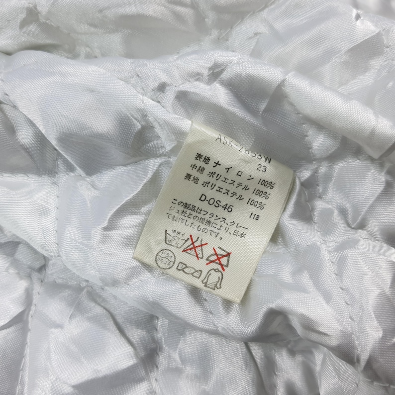 May include: Close-up of a white quilted fabric with a tag. The tag has Japanese text and reads "ASK-2653W 23". The tag also includes fabric composition details: 100% nylon, 100% polyester, and 100% polyester. Additional text on the tag includes "D-OS-46 118".
