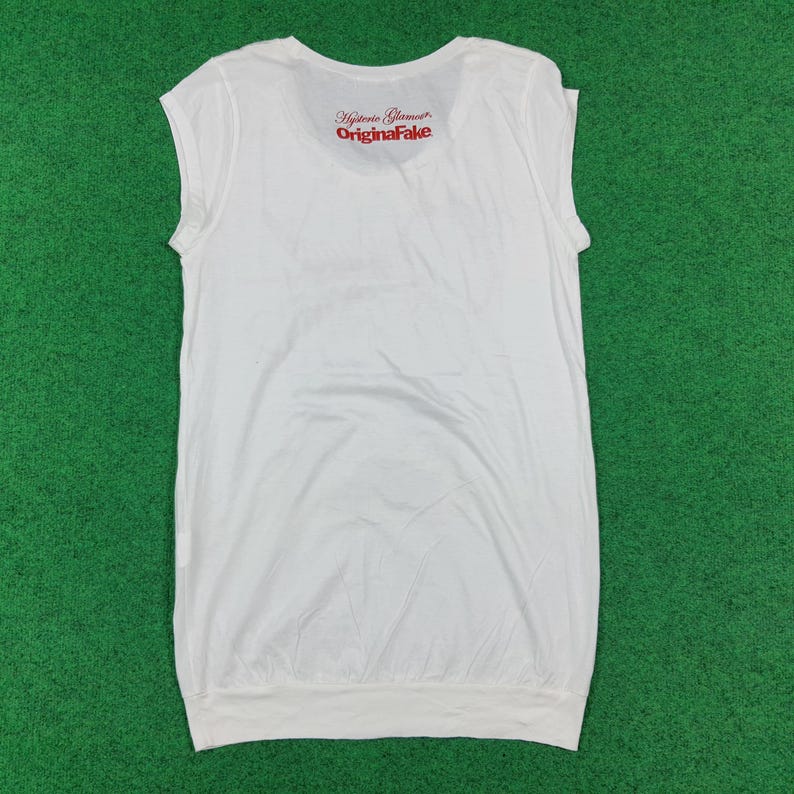 Puede incluir: Una camiseta blanca sin mangas con cuello redondo y un dobladillo ancho. En la parte superior de la espalda lleva impreso en rojo el texto Hysteric Glamour y OriginaFake. La prenda presenta un dise&ntilde;o sencillo y limpio con acabado liso.