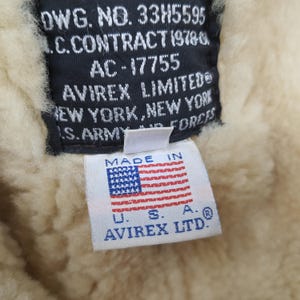 Puede incluir: Primer plano de una etiqueta de una chaqueta Avirex Limited. La etiqueta es blanca con el texto "MADE IN" encima de una peque&ntilde;a bandera americana. Debajo de la bandera est&aacute; el texto "U.S.A. AVIREX LTD.". El fondo es un material cremoso y difuso.