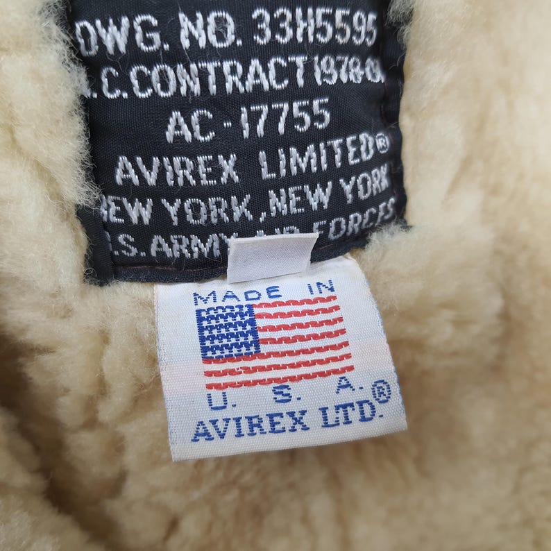 Puede incluir: Primer plano de una etiqueta de una chaqueta Avirex Limited. La etiqueta es blanca con el texto "MADE IN" encima de una peque&ntilde;a bandera americana. Debajo de la bandera est&aacute; el texto "U.S.A. AVIREX LTD.". El fondo es un material cremoso y difuso.