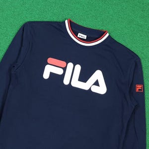 Puede incluir: Sudadera azul marino de manga larga con el logotipo blanco "FILA" en el pecho. El cuello tiene un dise&ntilde;o de rayas rojas y blancas. El logotipo de Fila tambi&eacute;n est&aacute; en la manga derecha.