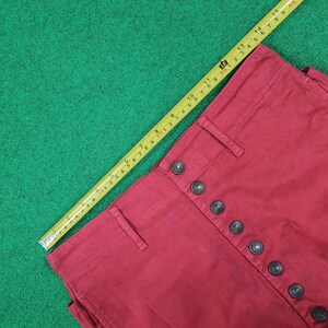 May include: Close-up of a pair of red pants with a button-up fly. The pants are measured with a yellow tape measure. The pants have a high waist and a series of black buttons.