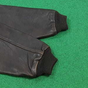 Puede incluir: Un par de mangas de chaqueta de cuero marr&oacute;n oscuro con pu&ntilde;os de punto acanalado negro en los extremos. Las mangas est&aacute;n construidas con costuras visibles a lo largo del cuero, mostrando una textura suave y un dise&ntilde;o estructurado.