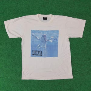 Puede incluir: Camiseta blanca con la portada del álbum "Nevermind" de Nirvana. El diseño muestra a un bebé nadando bajo el agua hacia un billete de dólar. El nombre de la banda y el título del álbum están impresos en negro.