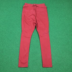 May include: A pair of red pants, viewed from the back, with a high waist and a belt detail. The pants are made of a solid-colored fabric and have a slim fit. The background is a solid green color.