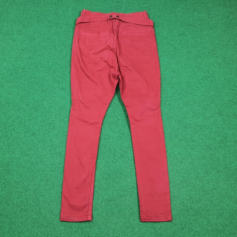 May include: A pair of red pants, viewed from the back, with a high waist and a belt detail. The pants are made of a solid-colored fabric and have a slim fit. The background is a solid green color.