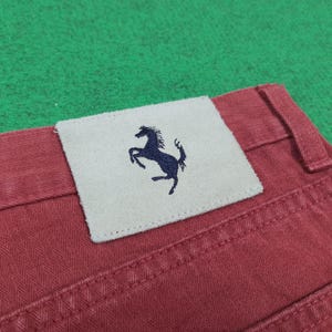 May include: Close-up of red denim pants with a stitched label featuring a navy blue prancing horse logo. The label is rectangular and made of a light-colored material. The pants have visible stitching.