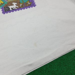 May include: White t-shirt with a colorful graphic of a stamp featuring a dog, a map, and other illustrations. The shirt has some light stains. The bottom of the shirt is visible, resting on a green surface.