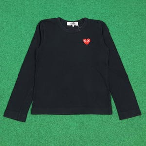 May include: Black long-sleeve shirt with a small red heart graphic with eyes on the chest. The shirt has the word "PLAY" printed on the upper chest. The shirt is made of a soft material.