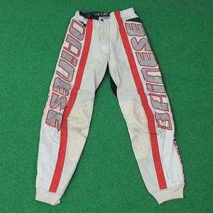 May include: White and gray motorcycle pants with red stripes and the word "DAINESE" printed vertically on the sides. The pants have knee padding and a black waistband. The pants are designed for motorcycle riding.