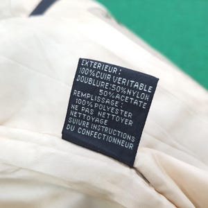 Puede incluir: Una etiqueta de cuidado rectangular negra cosida sobre un forro de tela color crema. La etiqueta presenta texto blanco que dice: EXTERIEUR: 100% CUIR VERITABLE, DOUBLURE: 50% NYLON 50% ACETATE, REMPLISSAGE: 100% POLYESTER, NE PAS NETTOYER, SUIVRE INSTRUCTIONS.