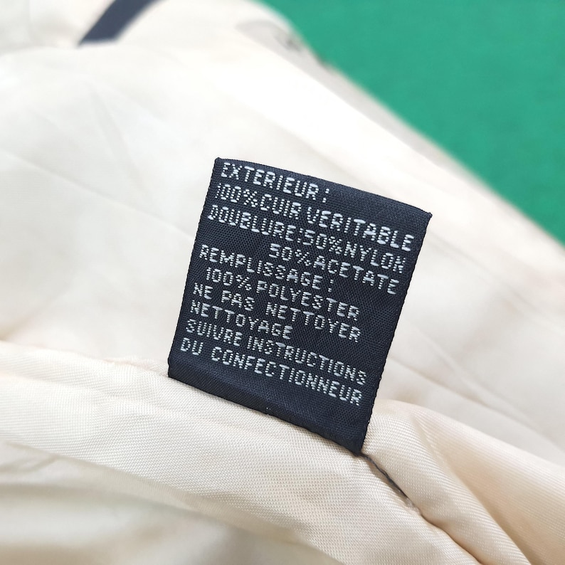 Puede incluir: Una etiqueta de cuidado rectangular negra cosida sobre un forro de tela color crema. La etiqueta presenta texto blanco que dice: EXTERIEUR: 100% CUIR VERITABLE, DOUBLURE: 50% NYLON 50% ACETATE, REMPLISSAGE: 100% POLYESTER, NE PAS NETTOYER, SUIVRE INSTRUCTIONS.