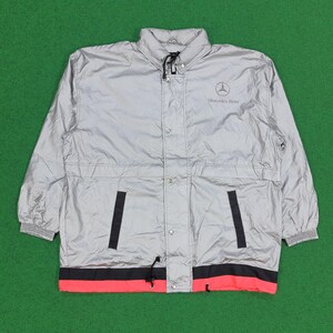 May include: A silver windbreaker jacket featuring a high collar, long sleeves with elastic cuffs, and a front snap-button closure. The chest displays a Mercedes-Benz logo and text. The hem includes a black and red horizontal stripe with drawstrings.