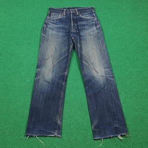K&ouml;nnte beinhalten: Verblasste blaue Jeans aus Denim mit Knopfleiste. Die Jeans haben ein klassisches F&uuml;nf-Taschen-Design und eine gerade Beinpassform. Der Denim hat ein abgenutztes Aussehen mit Ausbleichen und Abnutzung. Der Saum ist ausgefranst. Die Jeans liegen flach auf einer gr&uuml;nen Oberfl&auml;che.