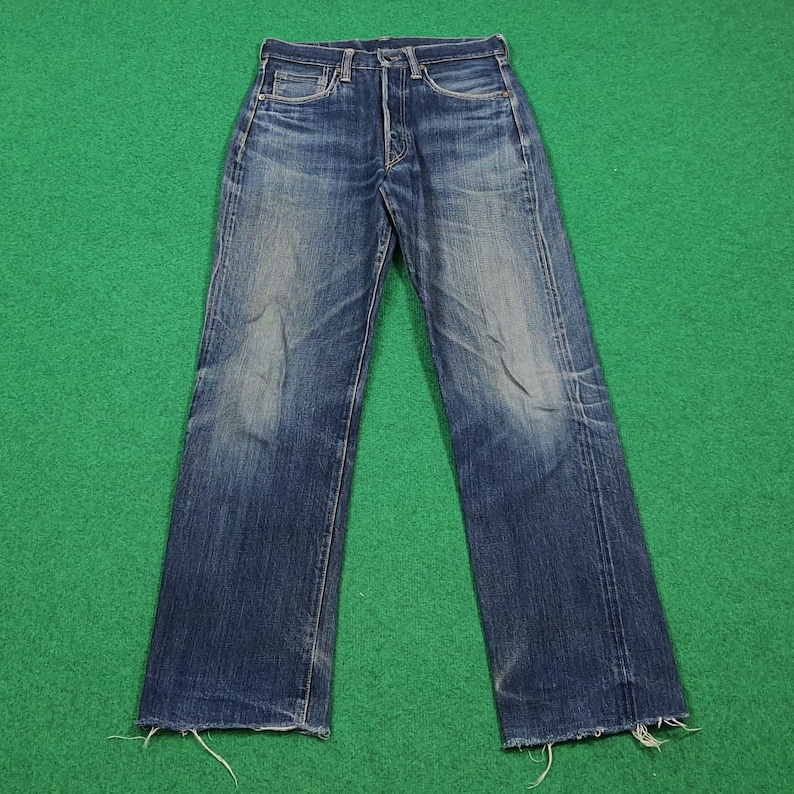 K&ouml;nnte beinhalten: Verblasste blaue Jeans aus Denim mit Knopfleiste. Die Jeans haben ein klassisches F&uuml;nf-Taschen-Design und eine gerade Beinpassform. Der Denim hat ein abgenutztes Aussehen mit Ausbleichen und Abnutzung. Der Saum ist ausgefranst. Die Jeans liegen flach auf einer gr&uuml;nen Oberfl&auml;che.